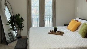 Un dormitorio con cama, ventilador de techo, ventana con persianas, mesita de noche con bandeja y una planta en maceta.