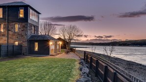 A modern house with a stone facade, a small attached structure with lights, and a view of a calm body of water at dusk. - 2 Bed in Inverness (oc-ca324) (Inverness)