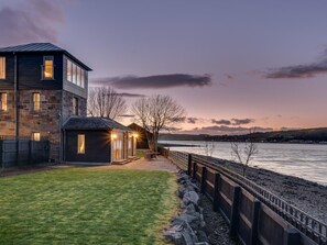 A modern house with a stone facade, a small attached structure with lights, and a view of a calm body of water at dusk. - 2 Bed in Inverness (oc-ca324) (Inverness)