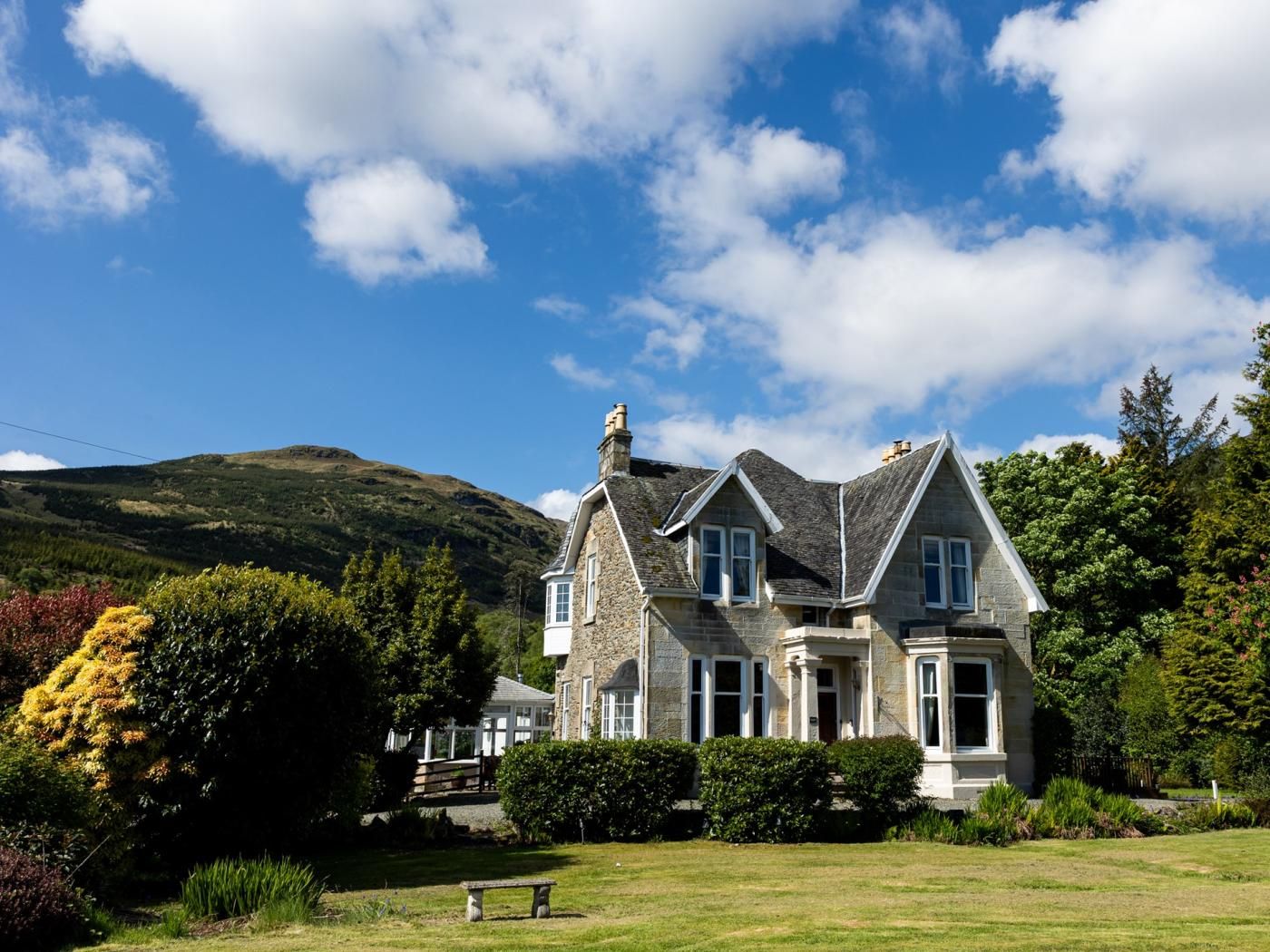 A stone house with a prominent chimney, surrounded by a well-maintained lawn and dense trees.