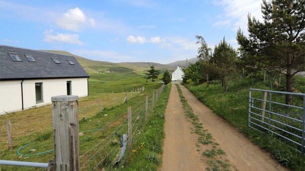 A rural road leading to a white house with a sloped roof, surrounded by greenery and hills. - 3 Bed in Isle Of Skye (oc-ca298) (Uig)