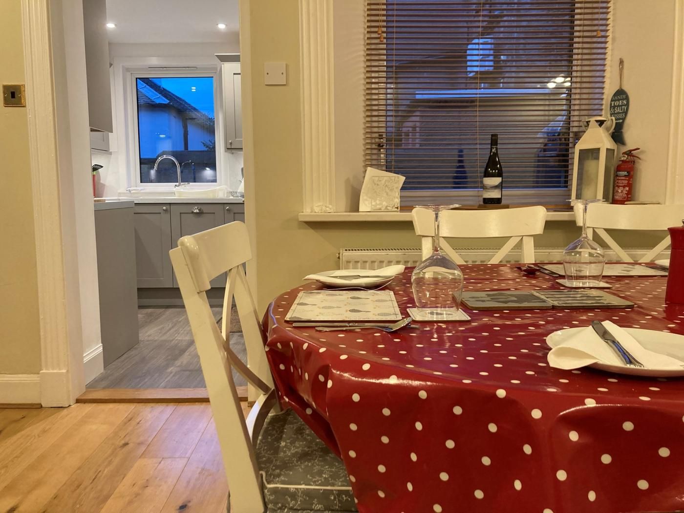 Comedor con mantel rojo, sillas blancas, una botella de vino y una ventana con persianas.