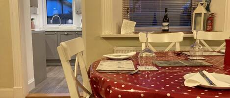 Comedor con mantel rojo, sillas blancas, una botella de vino y una ventana con persianas.