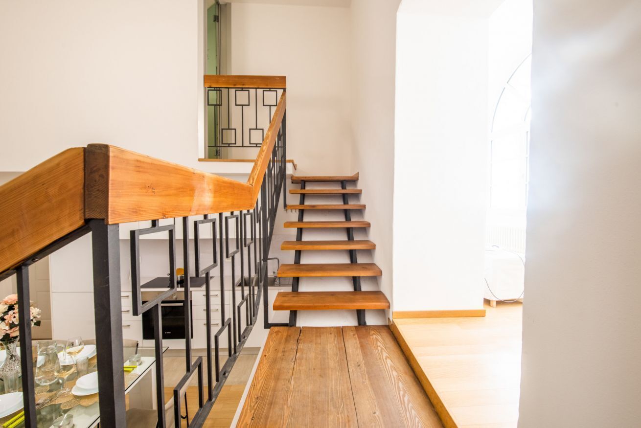 Una escalera de madera con barandilla de metal que lleva a una habitación con pared blanca y una ventana.