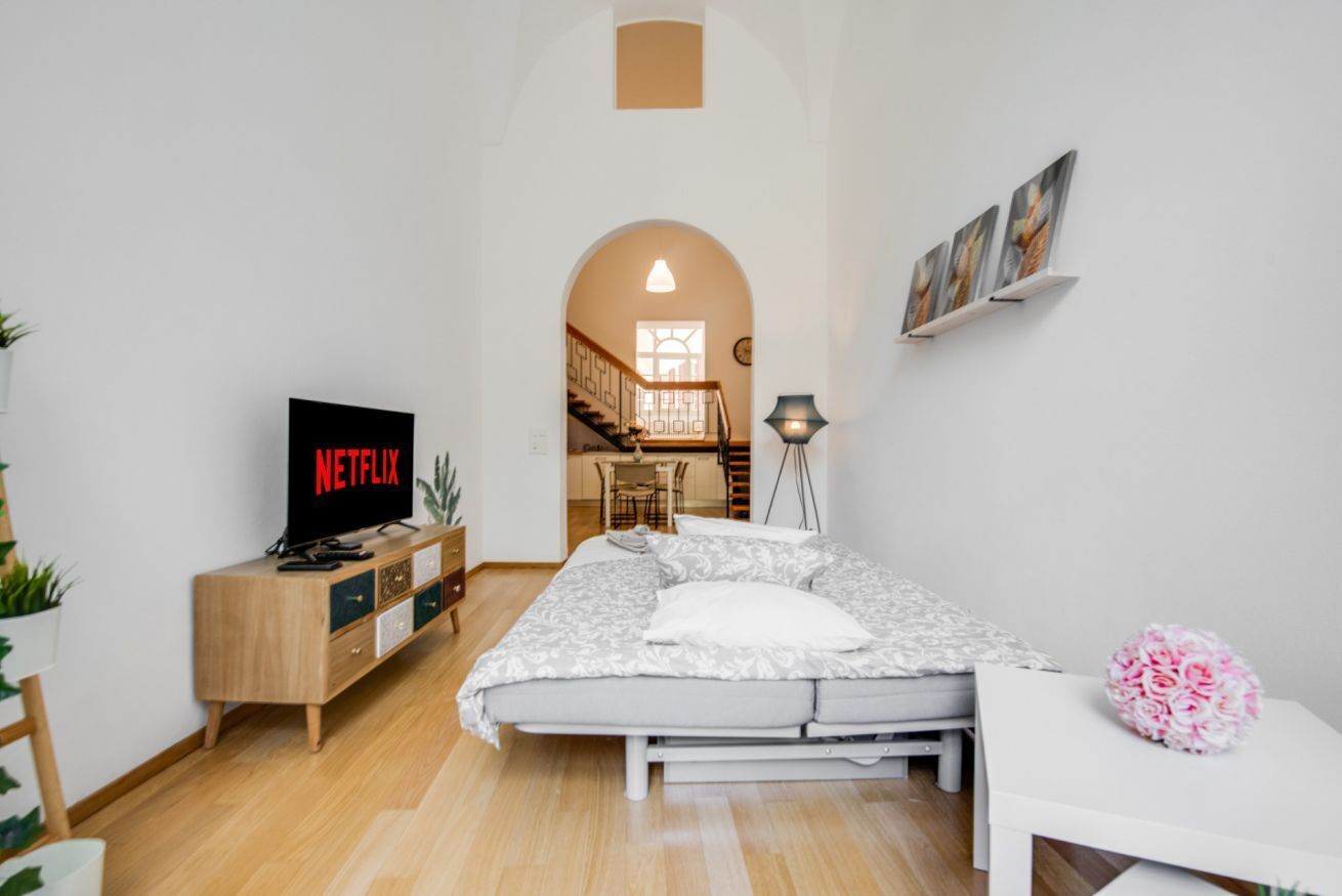 Un dormitorio con una cama, un televisor que muestra 'NETFLIX', una mesita de noche, una lámpara y vistas de una escalera que conduce a un comedor.