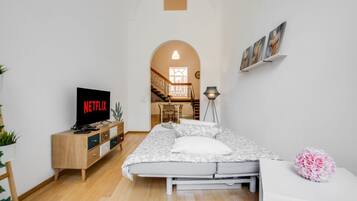 Un dormitorio con una cama, un televisor que muestra 'NETFLIX', una mesita de noche, una lámpara y vistas de una escalera que conduce a un comedor.