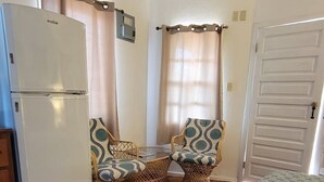 Una habitación pequeña con cama, refrigerador, dos sillones de mimbre, una mesa y una puerta.