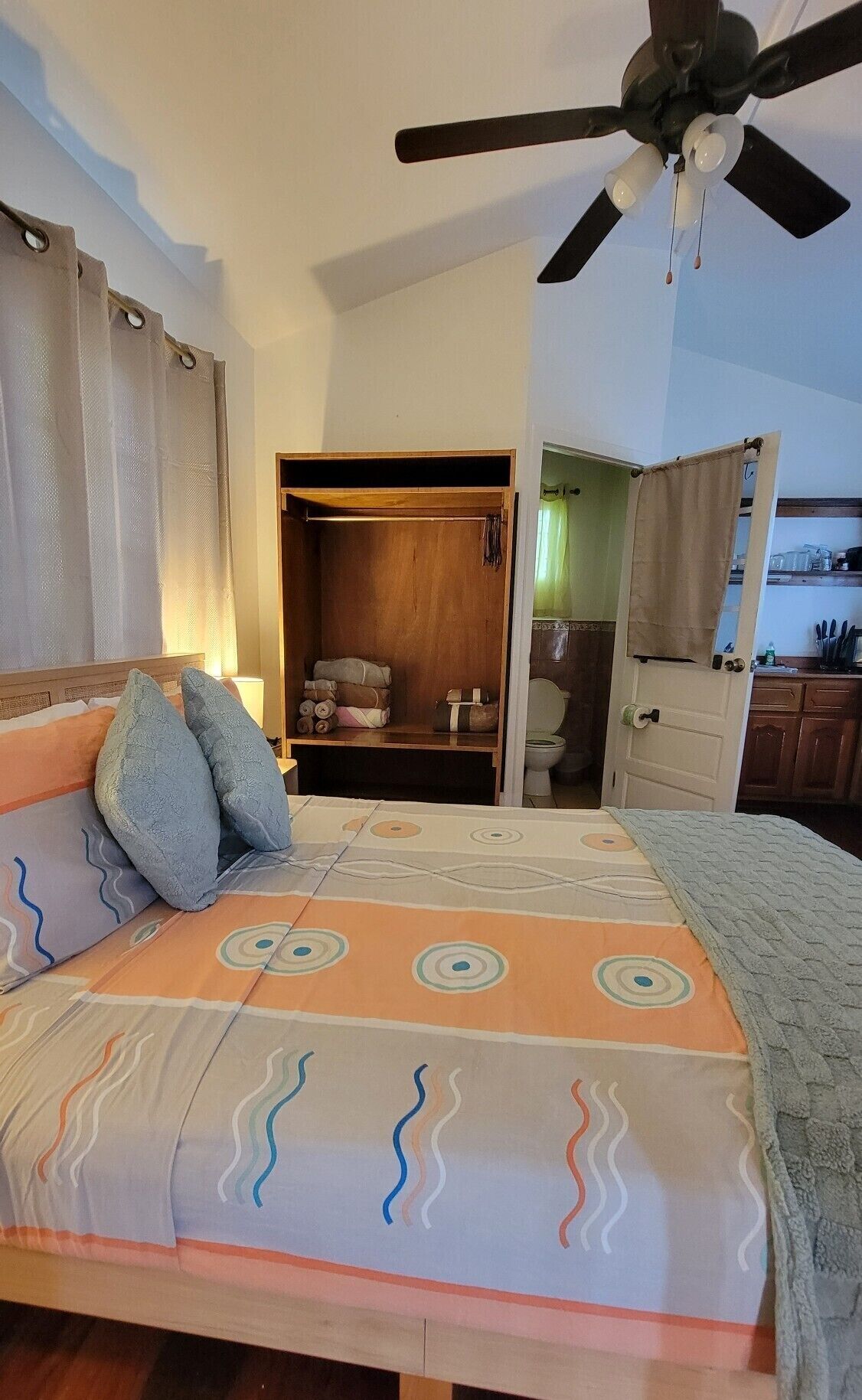 A bedroom with a bed, a ceiling fan, a wooden wardrobe, and a bathroom visible through an open door.