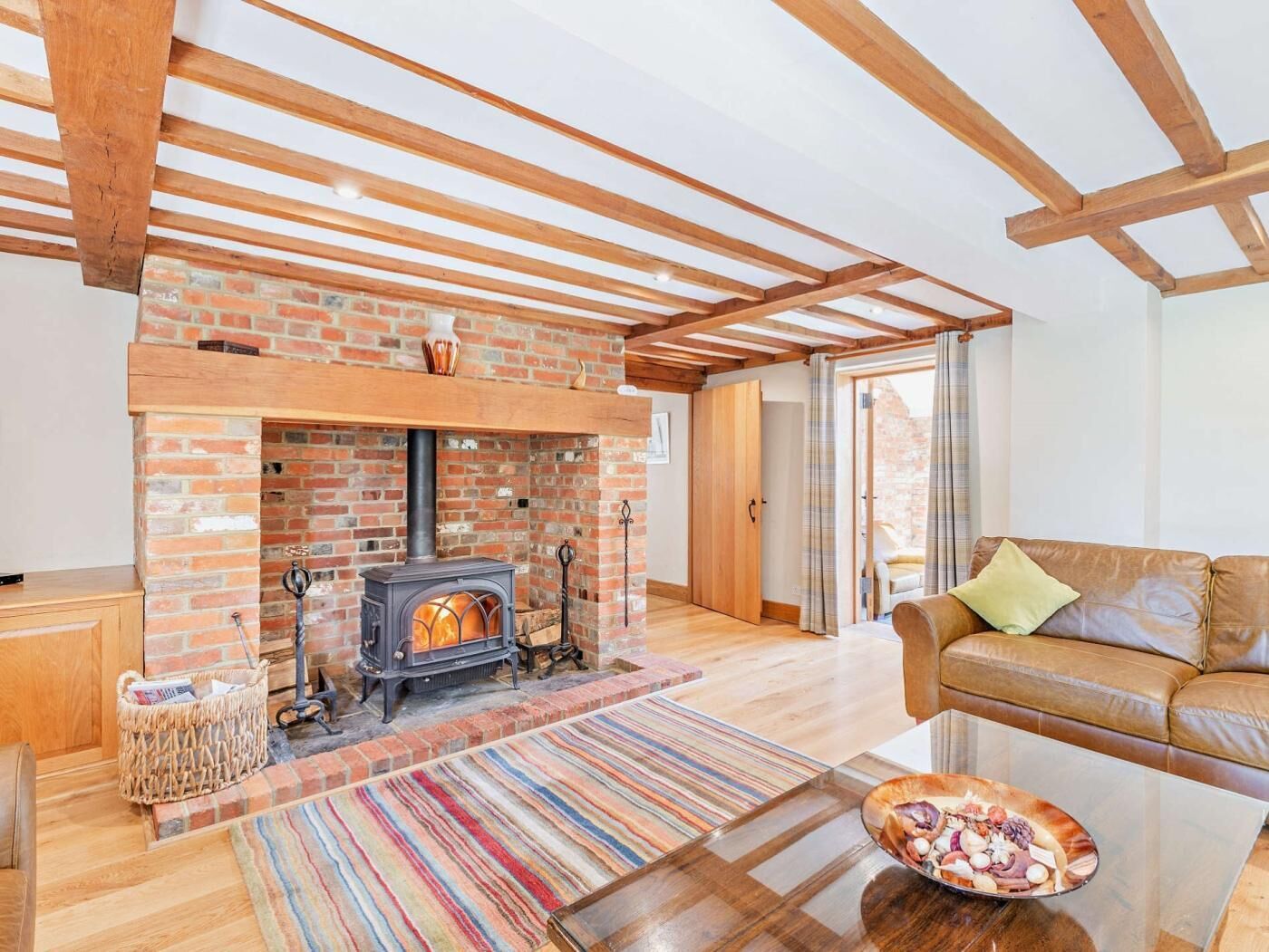 A living room with a brick fireplace, wooden beams, a leather sofa, and a colorful striped rug.