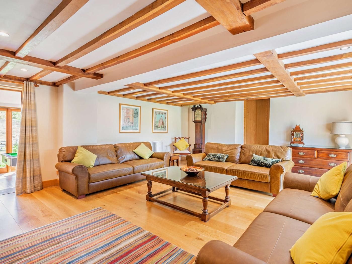 Ein geräumiges Wohnzimmer mit Holzbalken, einem braunen Ledersofa, einem Glastisch und einem Streifen Teppich.