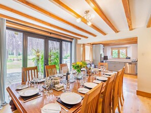 Een eetruimte met een lange houten tafel gedekt voor een maaltijd, grote ramen met uitzicht op groen, en een keuken op de achtergrond.