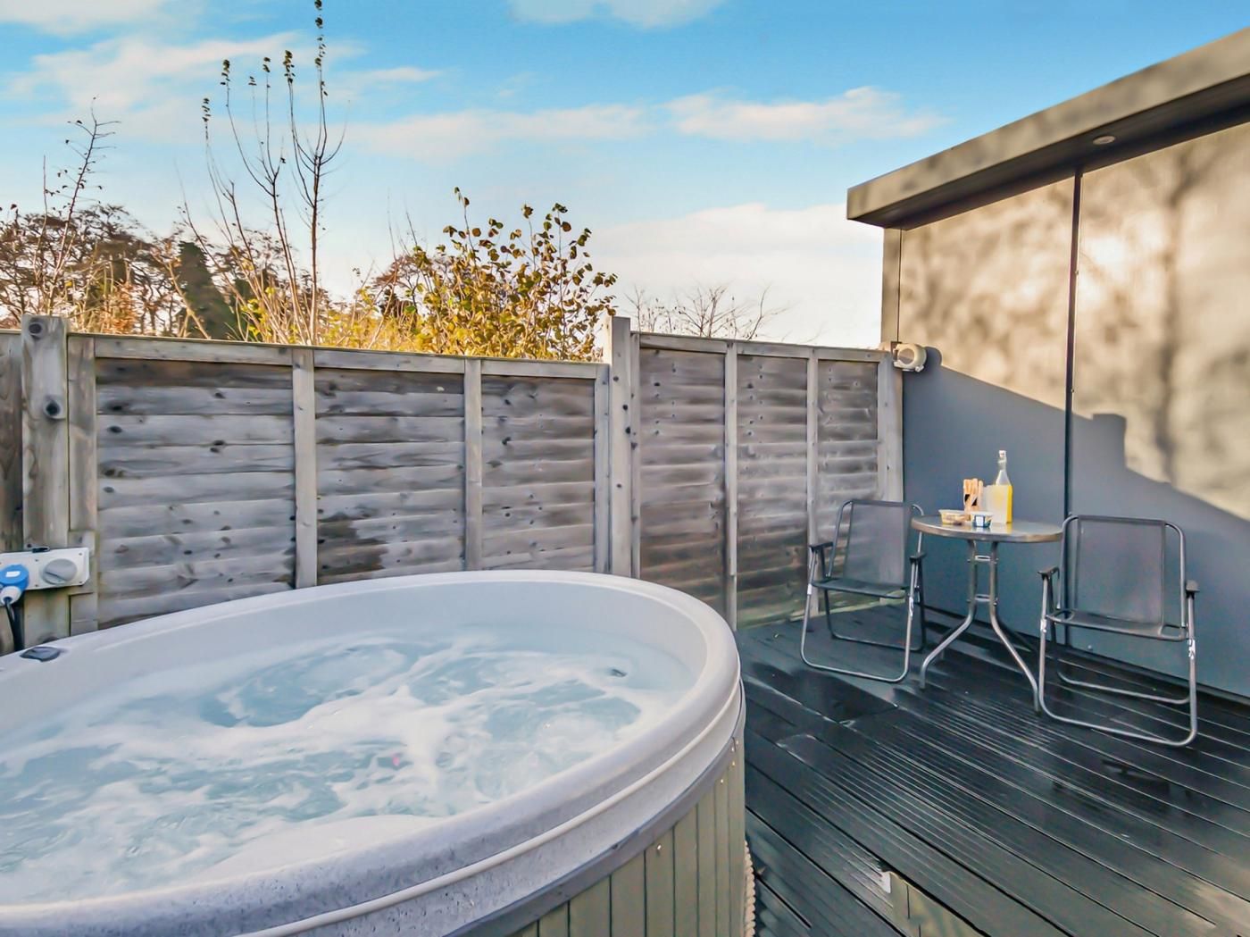 A hot tub on a wooden deck with a table and chairs, surrounded by a wooden fence and vegetation.