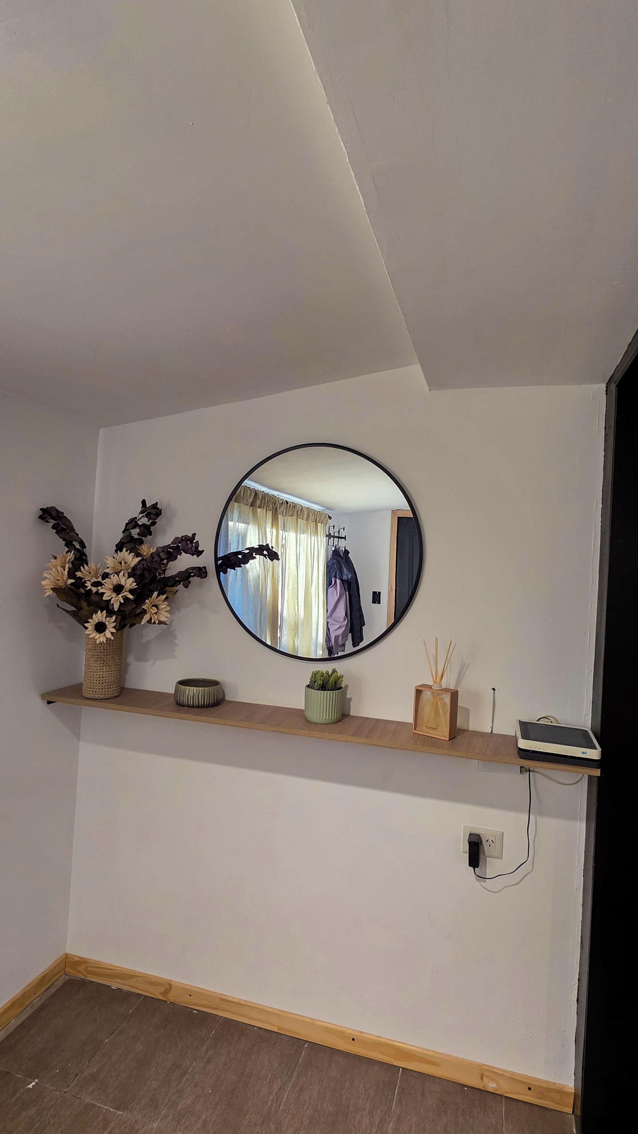 Una habitación con estante de madera, un espejo, un florero con flores secas, una planta en maceta y una pequeña mesa con un dispositivo.