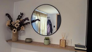 A room with a wooden shelf, a mirror, a vase with dried flowers, a potted plant, and a small table with a device.