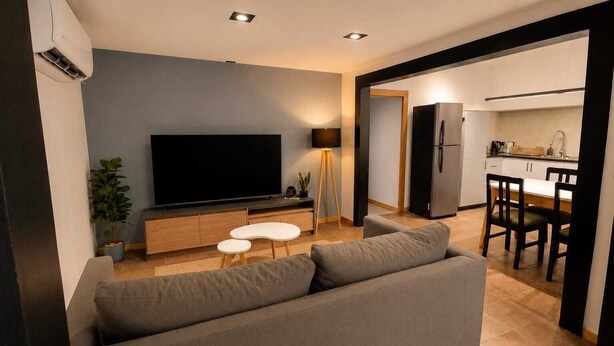 A modern living room with a large flat-screen TV, a grey sofa, a small white coffee table, and a dining area with a kitchen in the background.