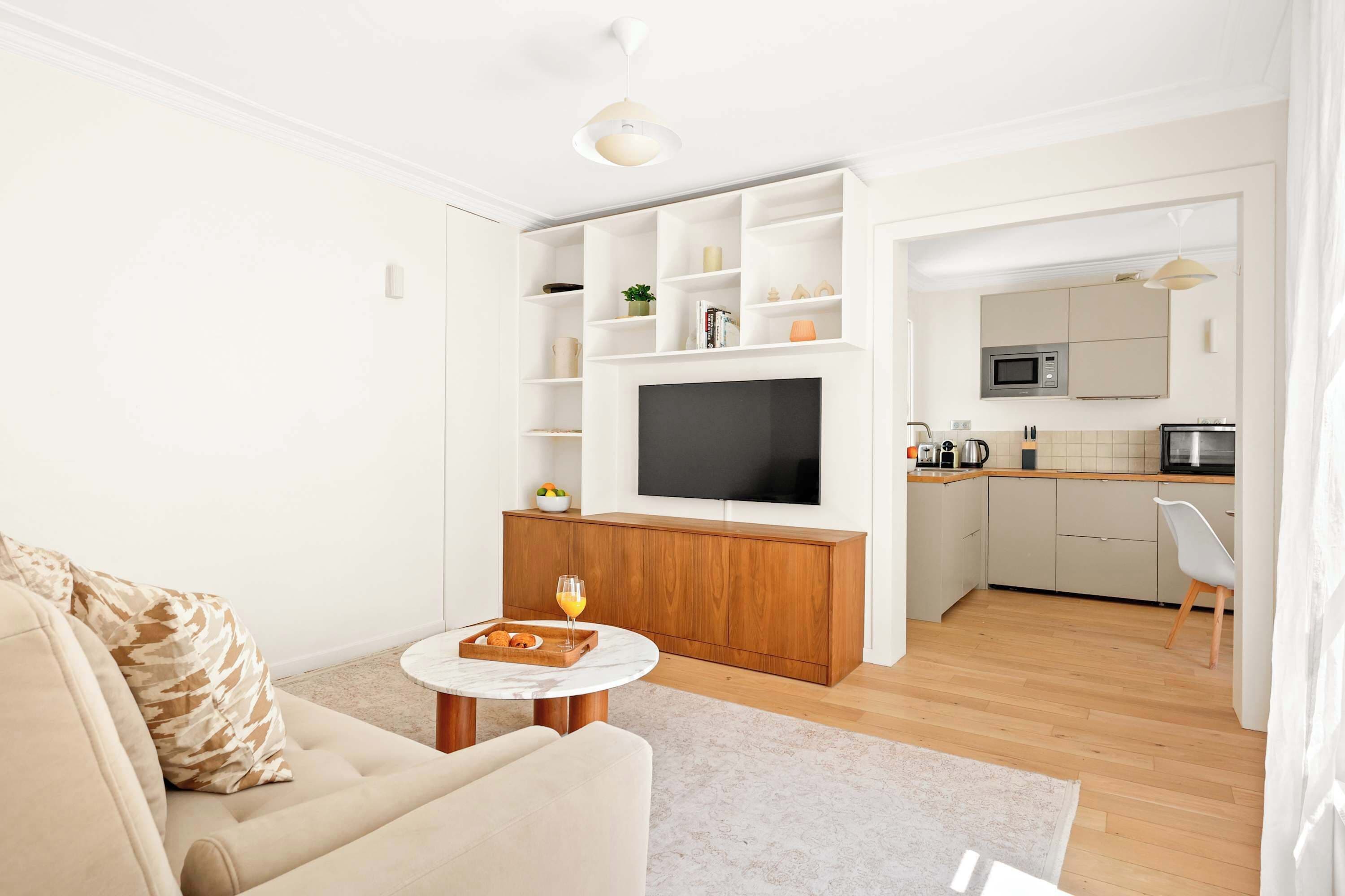 Un salon moderne avec un canapé, une table basse et un téléviseur. On peut apercevoir une cuisine équipée d’un four à micro-ondes et de placards à travers une porte ouverte.