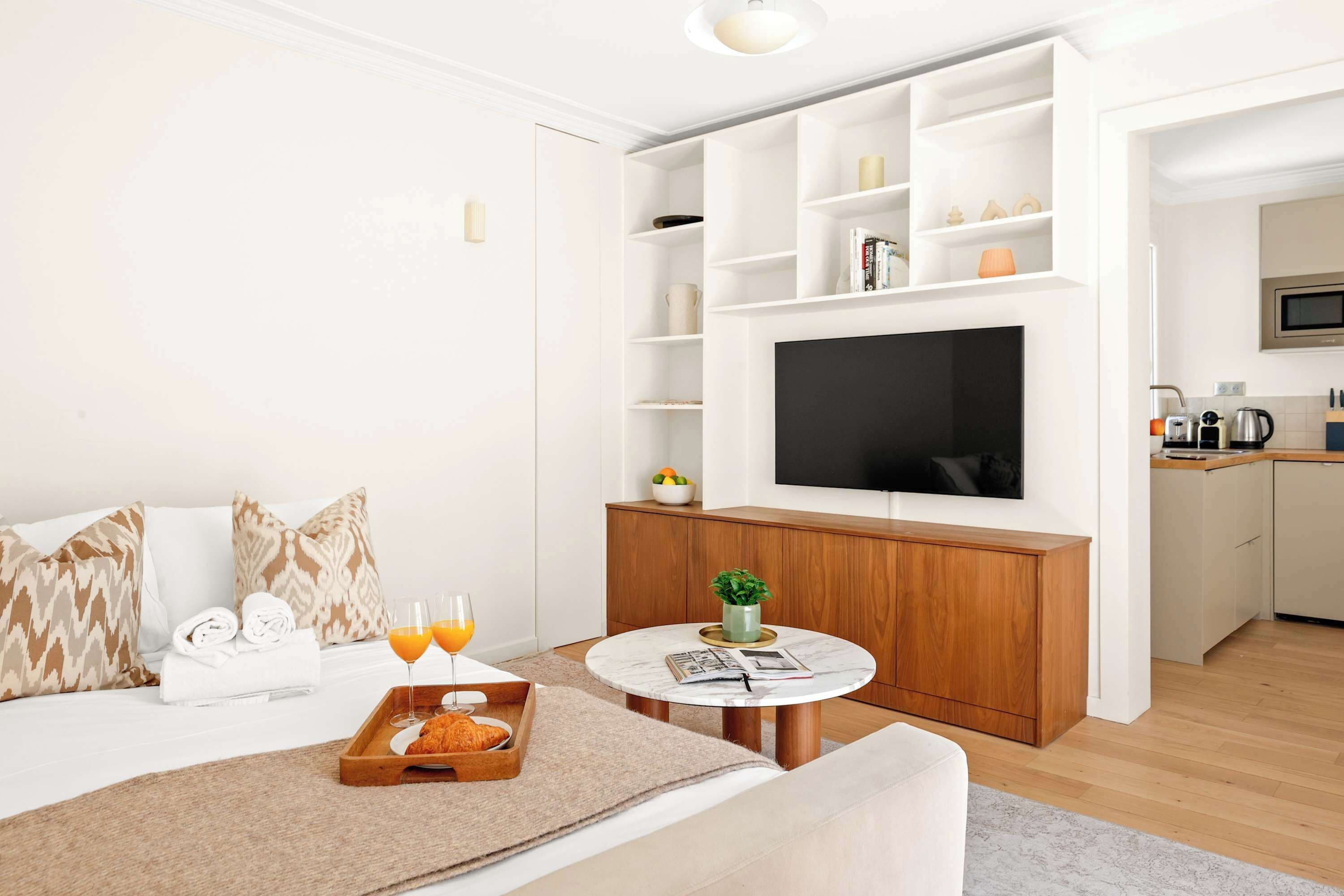 A hotel room with a bed, breakfast tray, two glasses of orange juice, a small round table with a magazine, a TV on a wooden cabinet, and a kitchen area with a microwave.