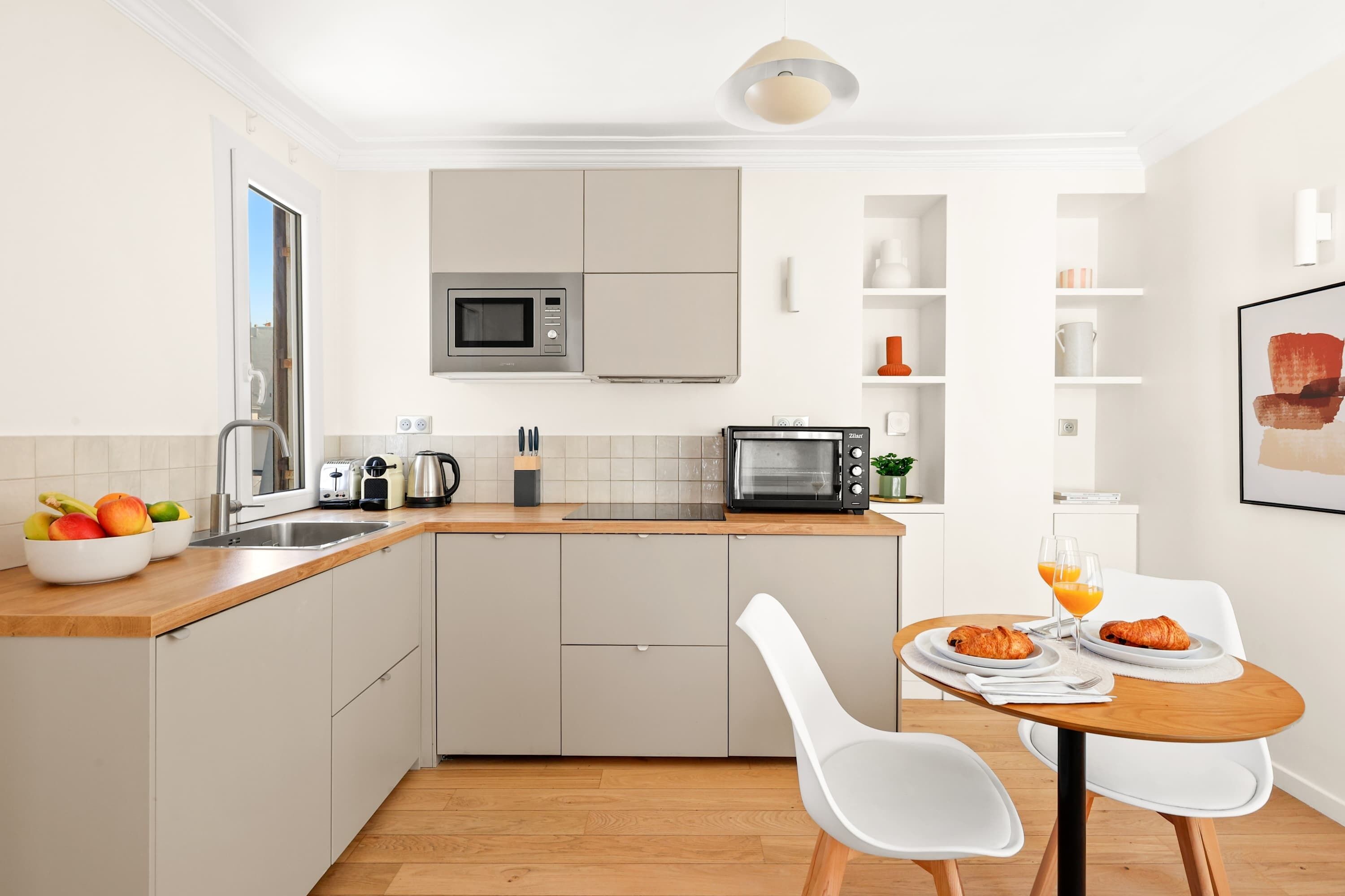 Een moderne keuken met een magnetron, broodrooster en spoelbak. Er staat een eettafel met stoelen en een schaal fruit.