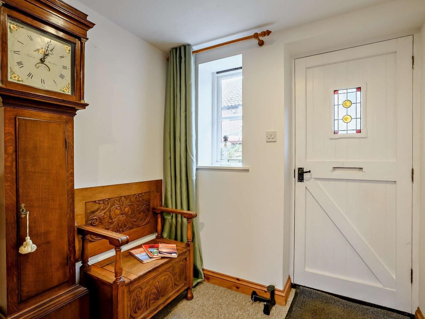 A room with a wooden bench, a clock, a door with a stained glass window, and a window with curtains.