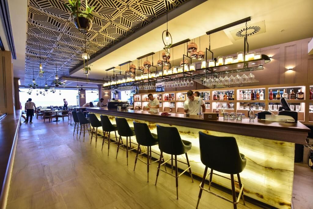 Un'area bar con un lungo bancone, diversi sgabelli da bar, luci a sospensione ed scaffali con bottiglie e bicchieri.