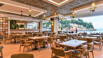 A restaurant with wooden tables and chairs, hanging plants, and a view of the sea.