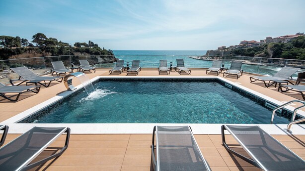 Una piscina con agua cristalina, rodeada de sillones de descanso, con vistas a una zona costera y edificios.