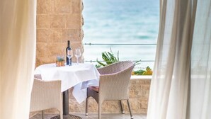 Una mesa con una botella y vasos, sillas de mimbre, vista al mar y una pared de piedra.