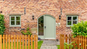 A brick house with a green door, two windows, a wooden fence, and a potted plant. - 3 Bed in Church Stretton (oc-76621) (Rushbury)