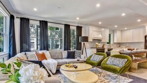 Una sala moderna con un sofá, dos sillones, una mesa de centro y un comedor, con una cocina al fondo.
