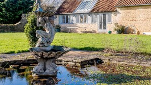 Ein Steinkunstwerk mit einer Figur in dynamischer Pose, ein Teich mit Seerosen und ein Steinhaus mit Ziegeldach und einem Wintergarten im Hintergrund.