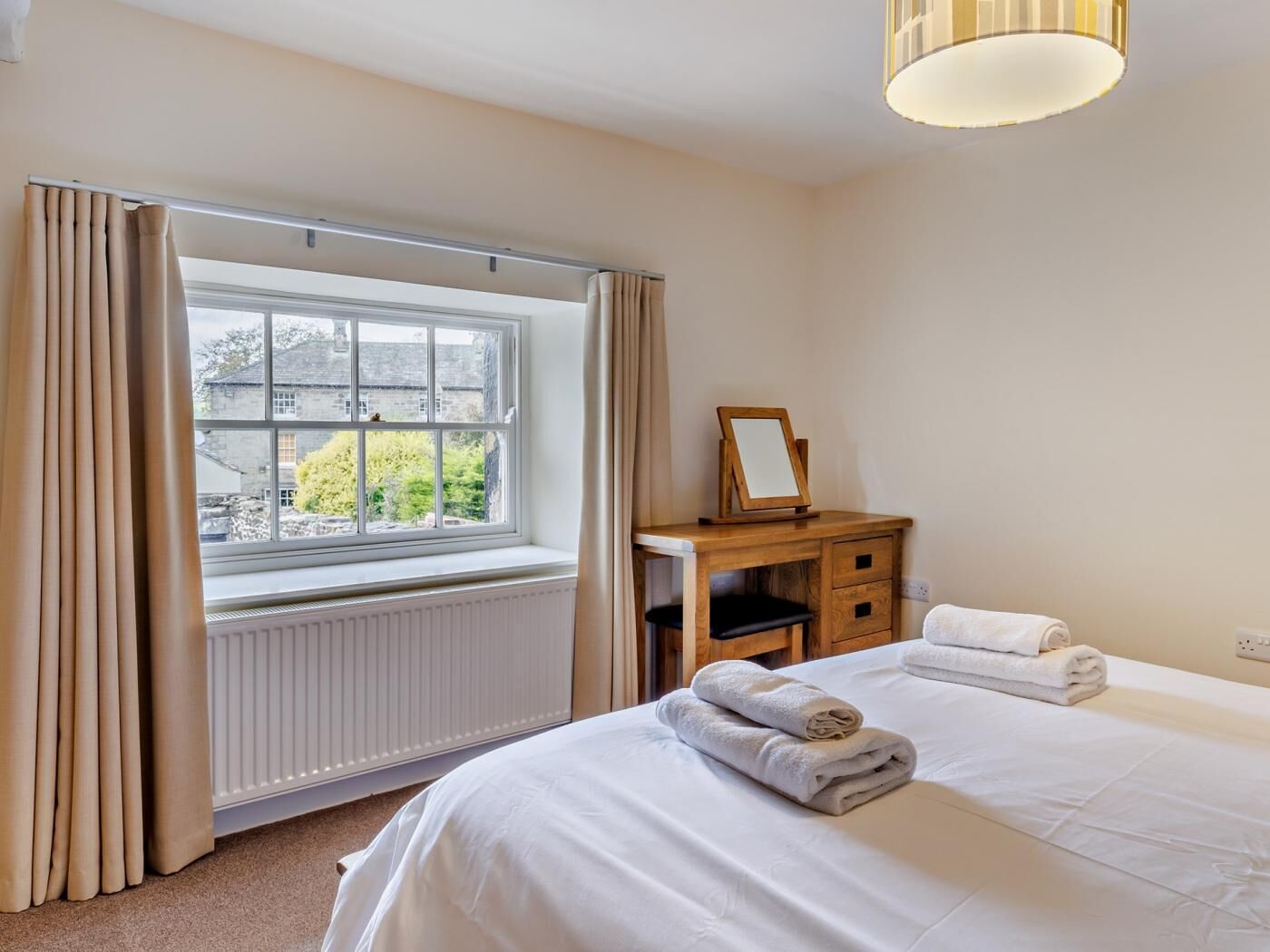 A hotel room with a large window, a wooden desk with a mirror, two beds with towels, and a radiator.