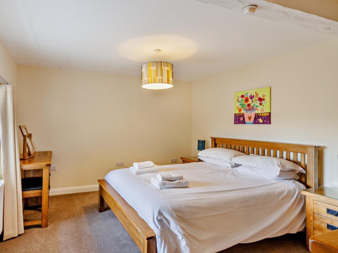 A bedroom with a wooden bed, a bedside table, a nightstand, a hanging lamp, and a colorful artwork on the wall.