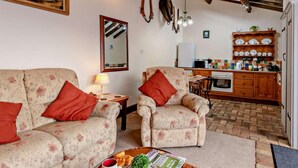 A living room with floral-patterned sofas, a coffee table with magazines and a plant, a kitchen with wooden cabinets and a tiled backsplash, and a rustic ceiling. - 2 Bed in King's Lynn (oc-77764) (Tattersett)
