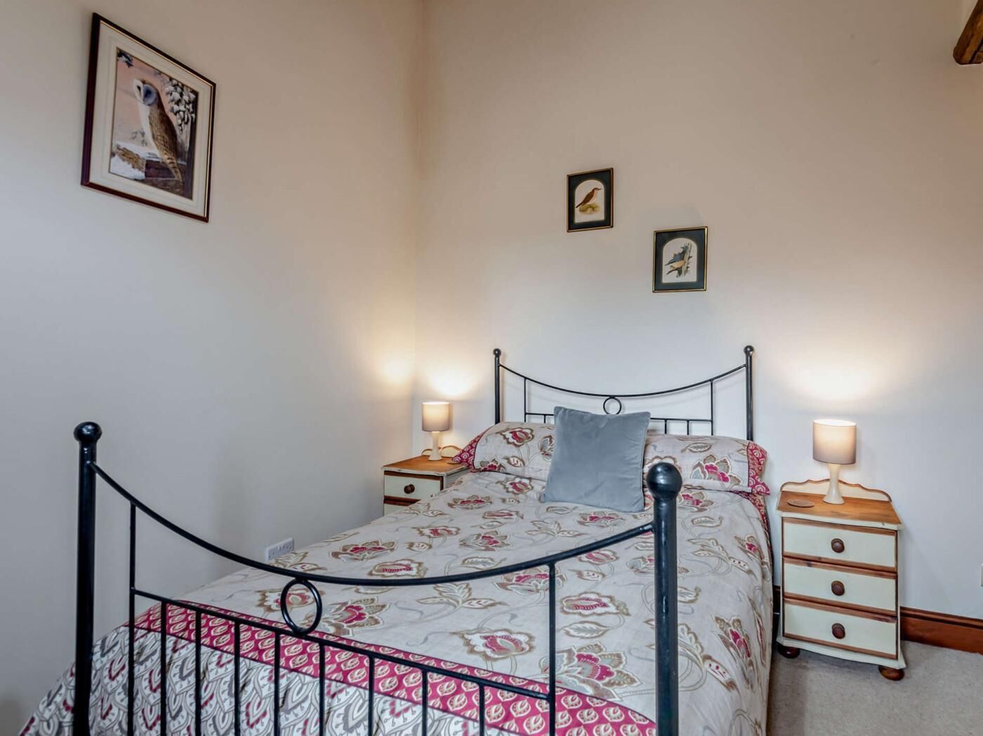 Quarto com estrutura de cama em metal, roupa de cama floral, duas mesas de cabeceira com abajures e quadros nas paredes.