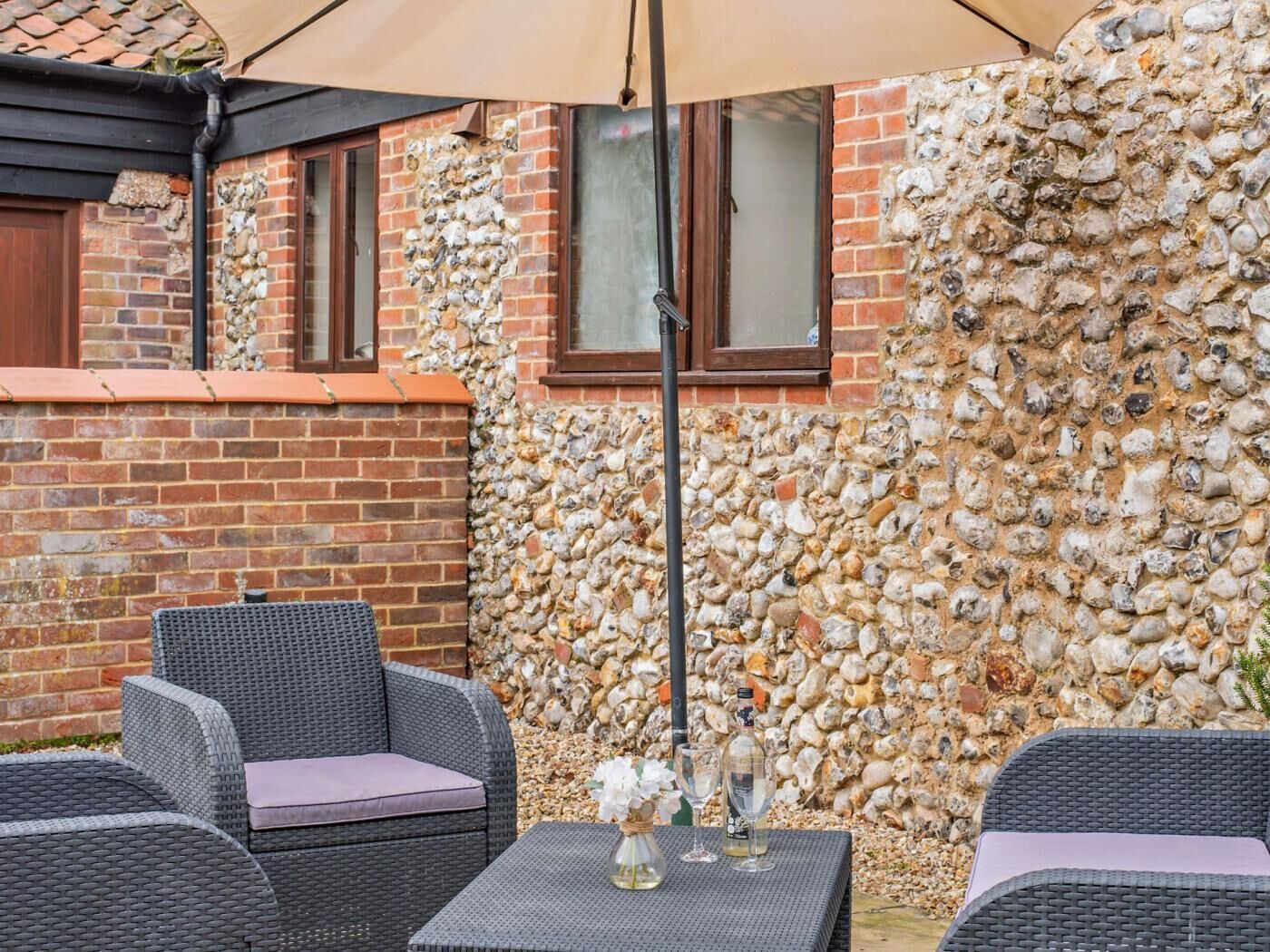 An outdoor seating area with wicker furniture, a table with a vase and glasses, and a stone wall backdrop.