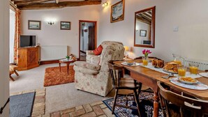 A cozy living room with a wooden ceiling, a television, a floral armchair, a dining table with breakfast, and a radiator. - 2 Bed in King's Lynn (oc-77764) (Tattersett)