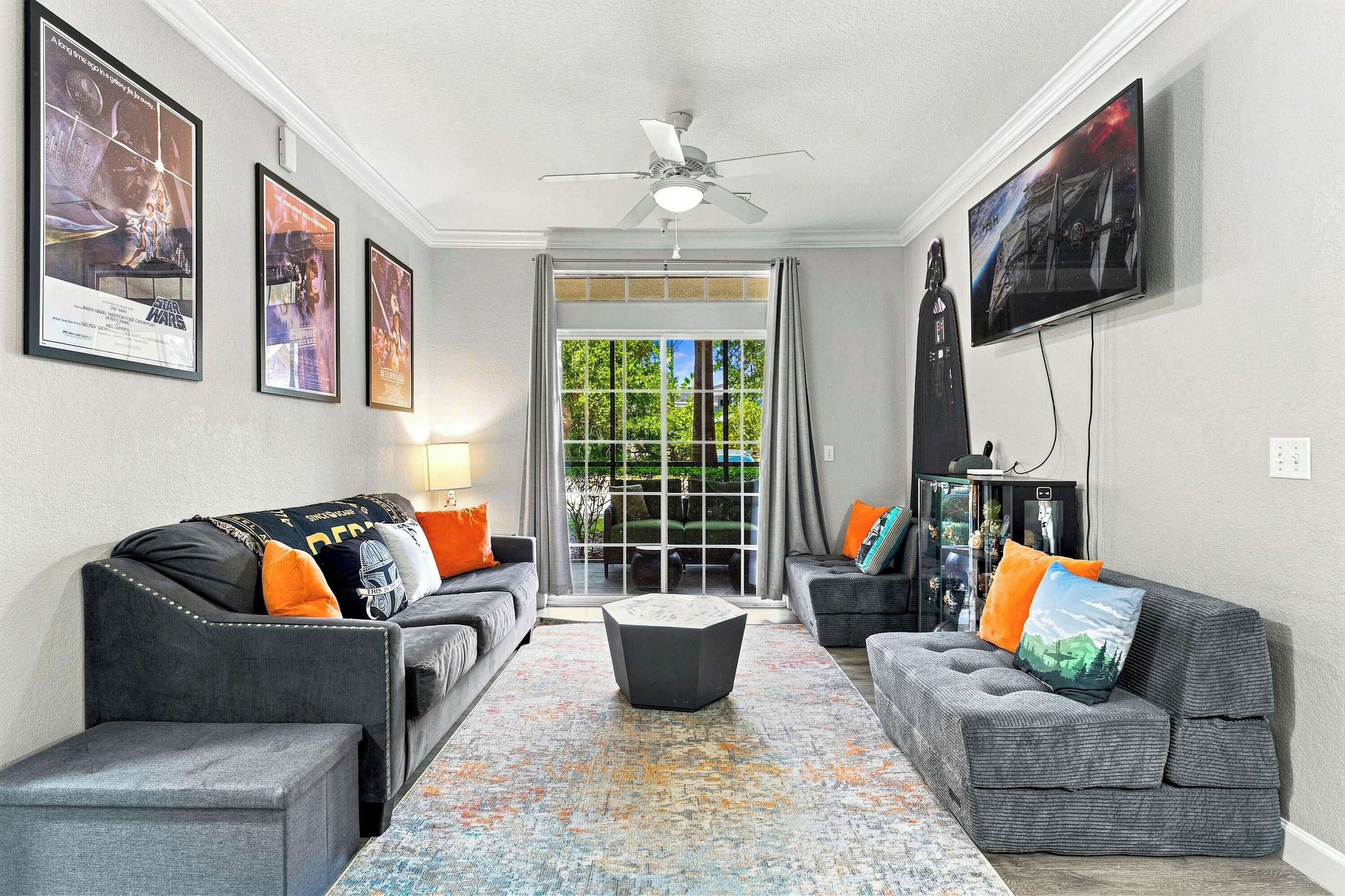 A living room with two grey sofas, a coffee table, and a ceiling fan. There are framed posters on the walls and a television mounted on the wall.