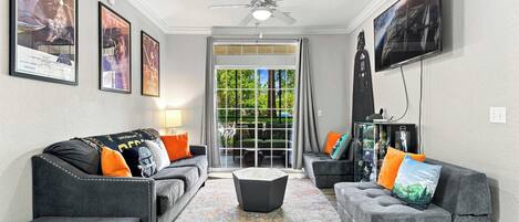 A living room with two grey sofas, a coffee table, and a ceiling fan. There are framed posters on the walls and a television mounted on the wall.