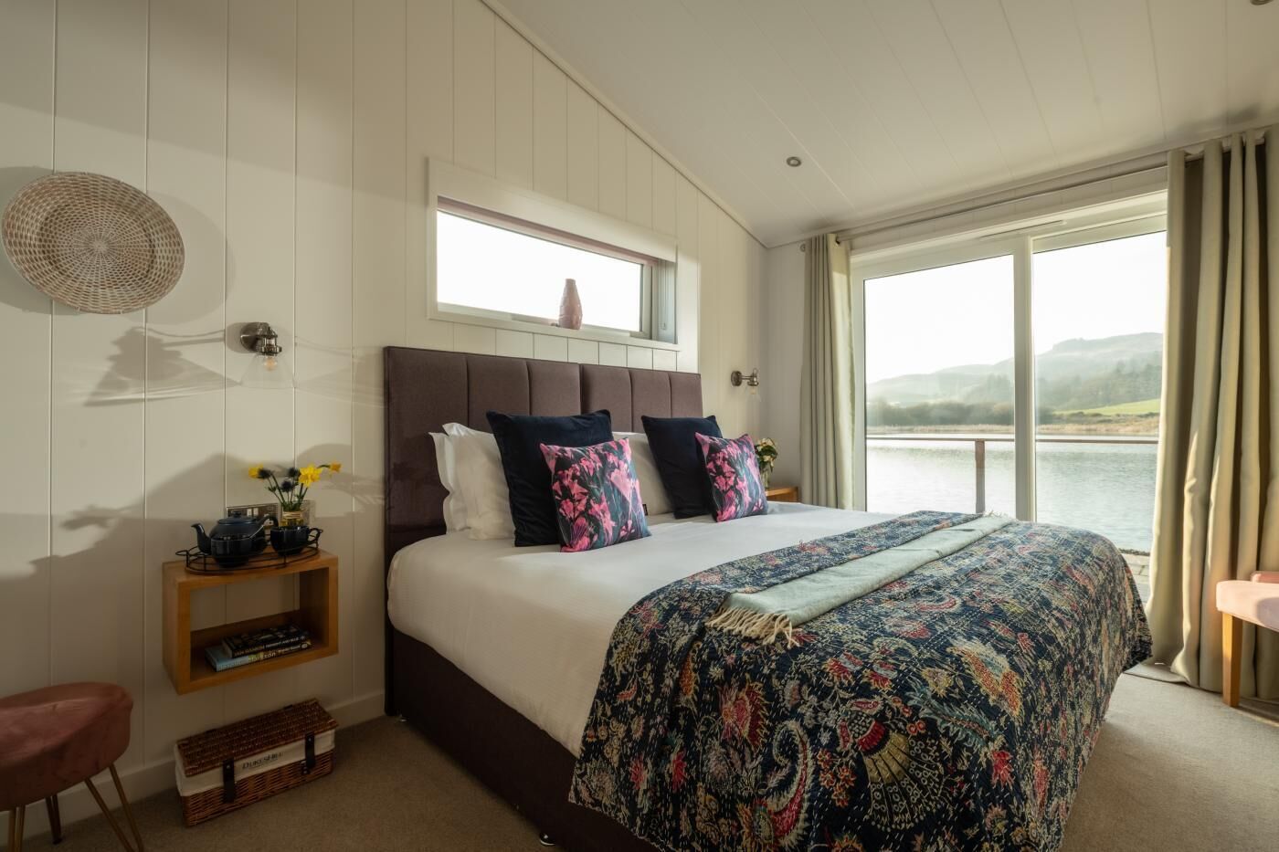 Un dormitorio con una cama grande, una ventana con vistas a un lago, una mesita de noche con una lámpara y un estante pequeño con libros.