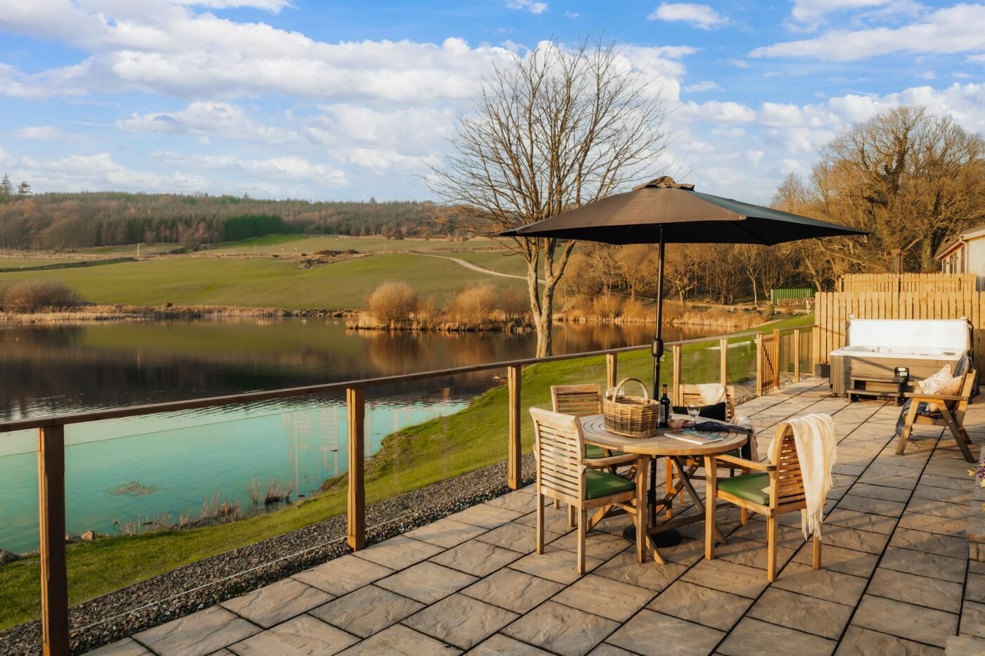 Une terrasse dotée de mobilier d’extérieur, donnant sur un lac, avec une clôture en bois et un ciel dégagé.