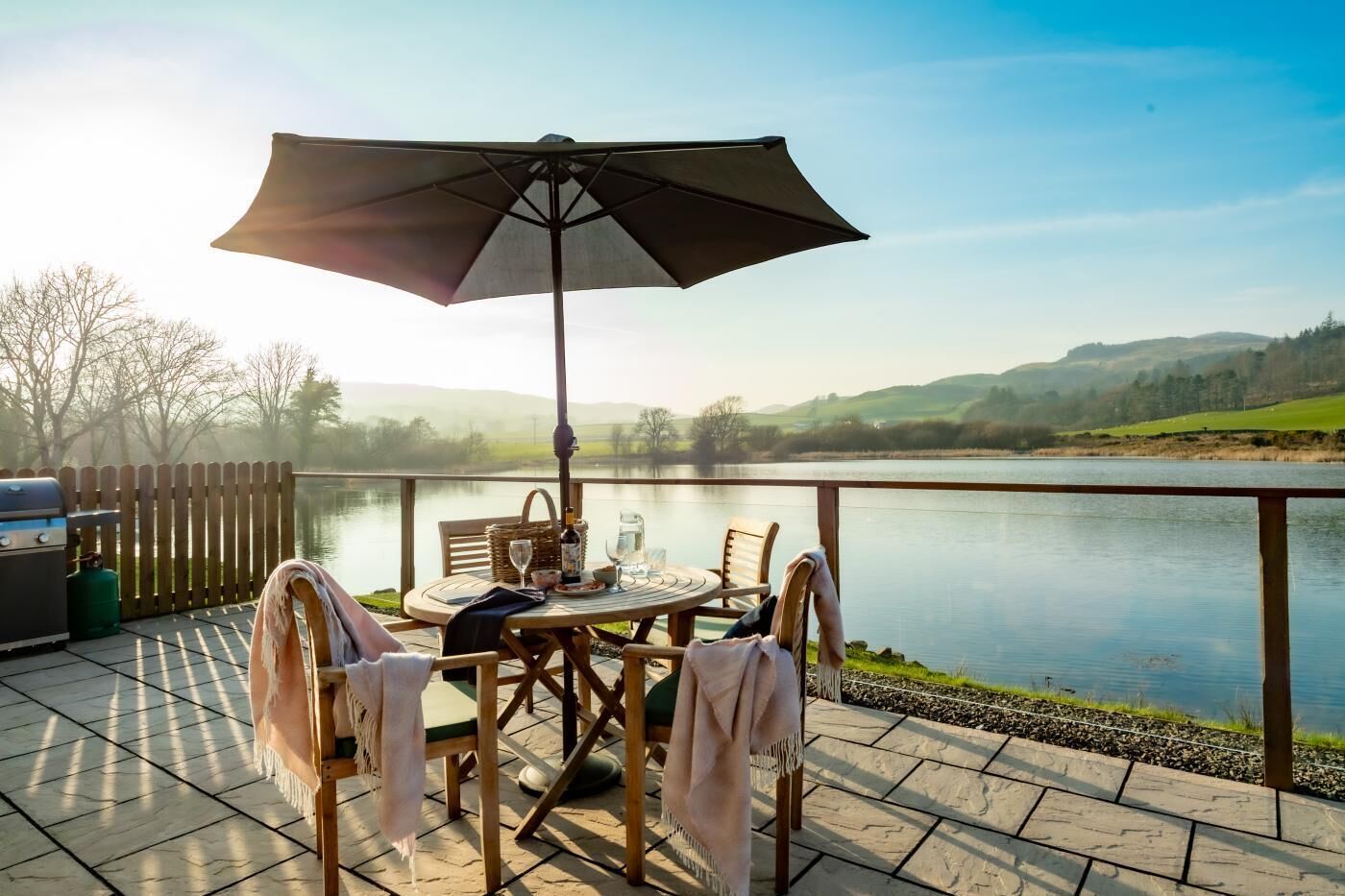 Eine Terrasse mit Tisch, Stühlen, einem Grill und Blick auf einen See und Hügel.
