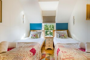 Quarto com duas camas, janela com persianas, mesa de cabeceira com abajur e aquecedor fixado na parede.