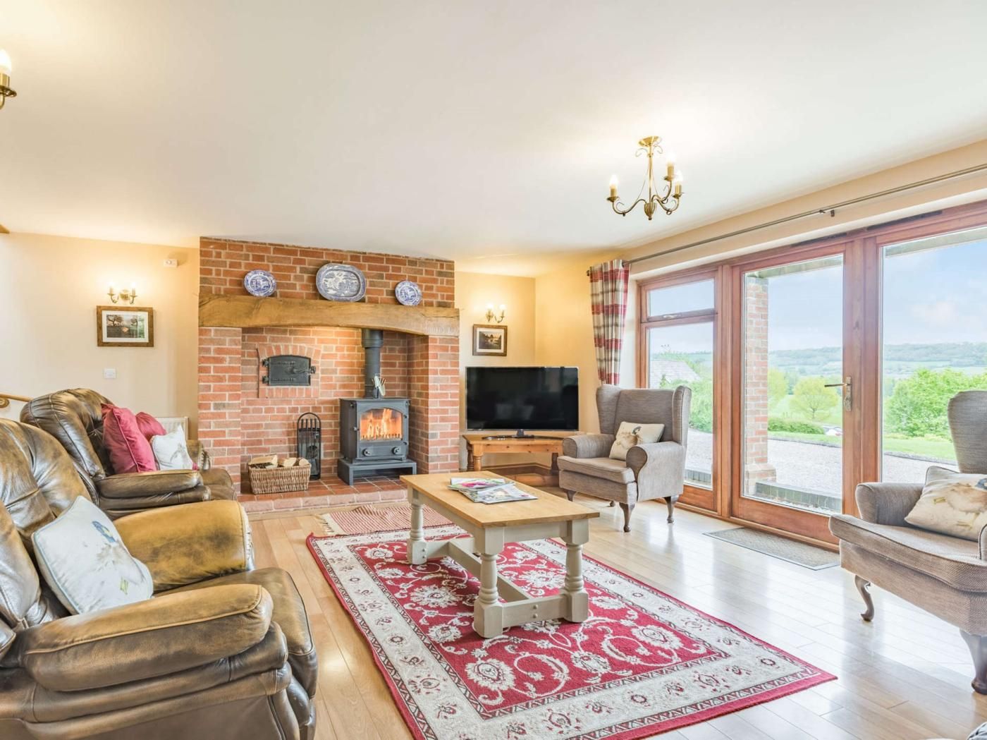 A cozy living room with a brick fireplace, leather sofas, a television, and a large window offering a view of the outdoors.