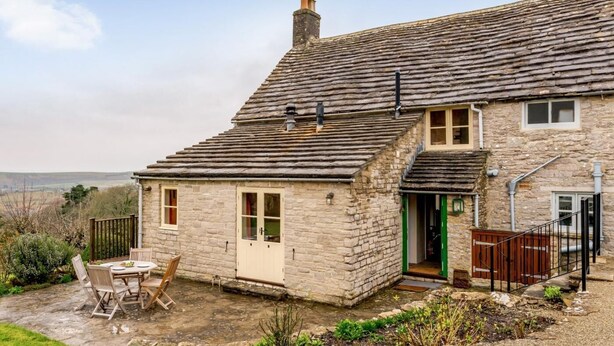 A stone house with a sloped roof, a small patio with a table and chairs, and a view of the countryside. - 4 Bed in Wareham (oc-78042) (Kingston)