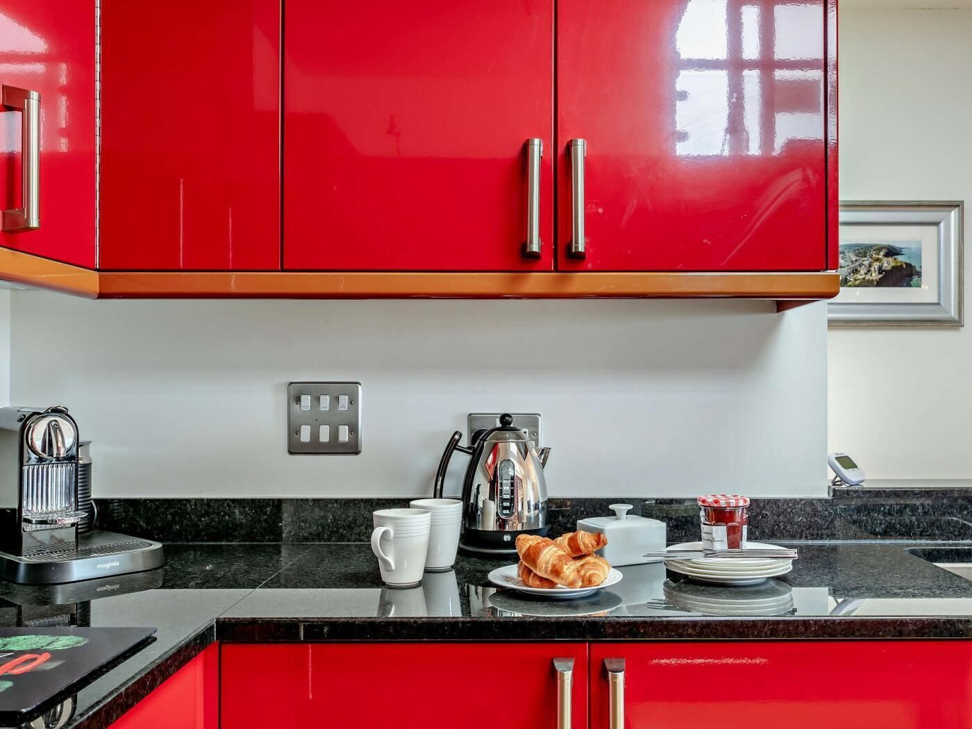 A kitchen with red cabinets, a coffee maker, a kettle, and a plate with croissants.