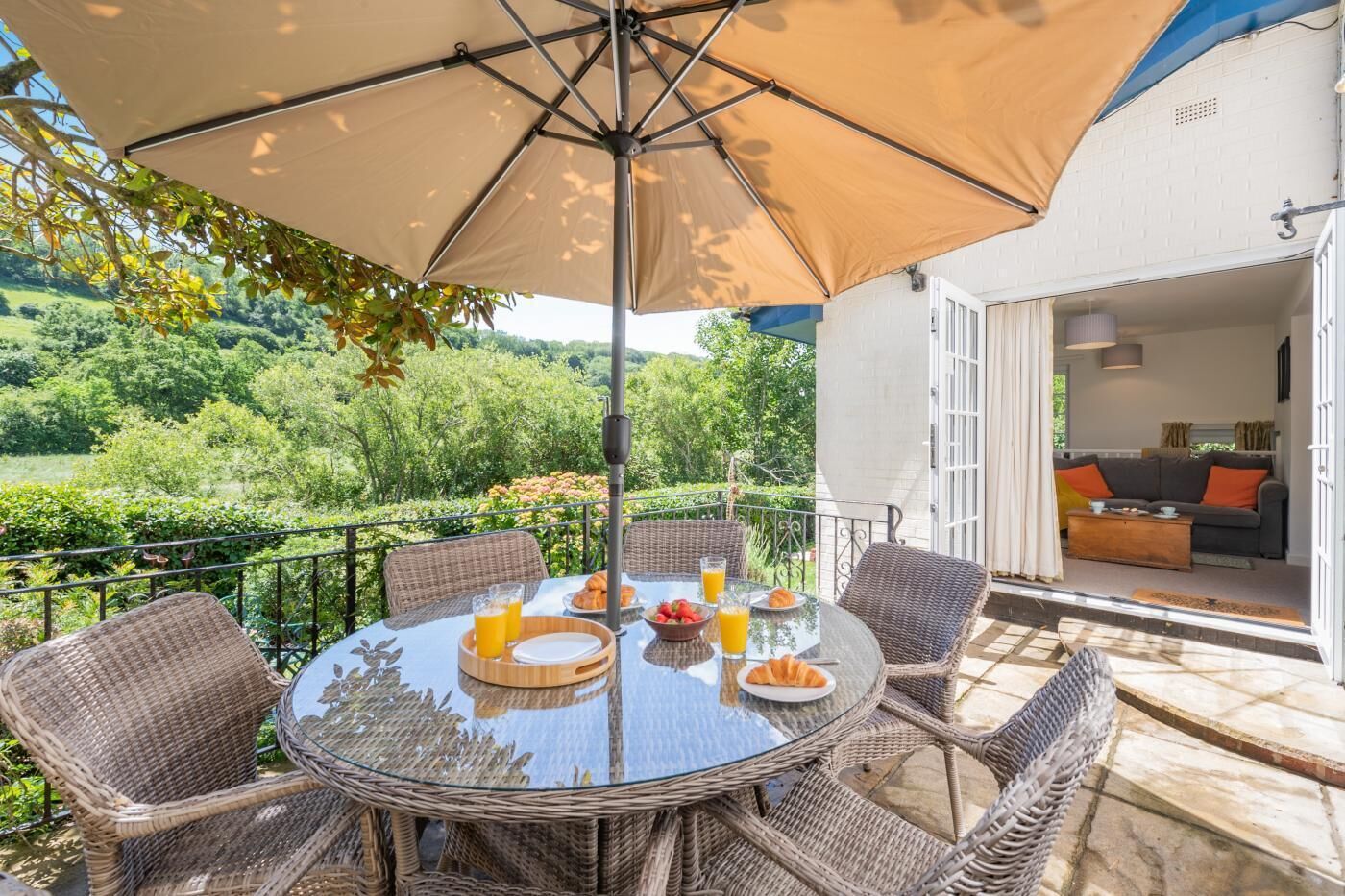 Ein runder Tisch mit Korbstühlen und einem großen Sonnenschirm, auf einem Balkon mit Blick auf einen üppigen Garten und Hügel.