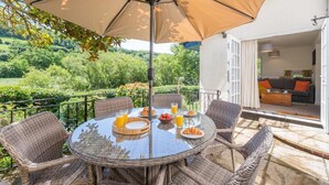 A round table with wicker chairs and a large umbrella, set on a balcony with a view of a lush garden and hills. - 3 Bed in Branscombe (oc-78002) (Branscombe)