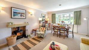 A cozy living room with a fireplace, a dining area, and a view of greenery through the window. - 3 Bed in Branscombe (oc-78002) (Branscombe)