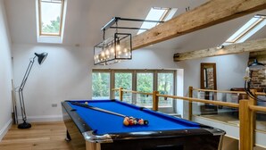 Ein Billardtisch mit Kugeln, einer Lampe und einem Billardqueue in einem Raum mit Holzbalken und großen Fenstern.