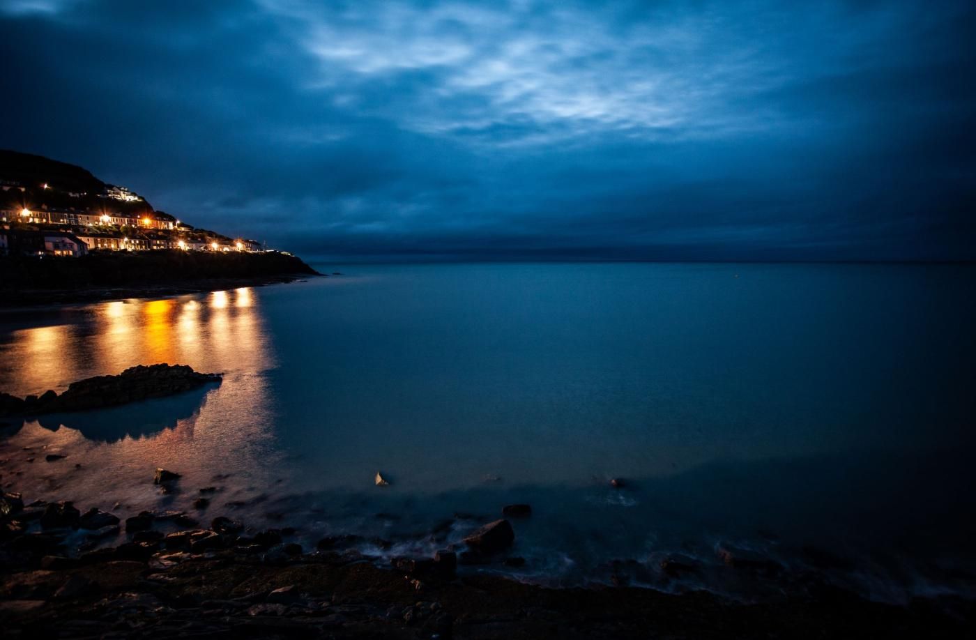 A coastal scene with a calm sea reflecting lights from a distant shoreline.